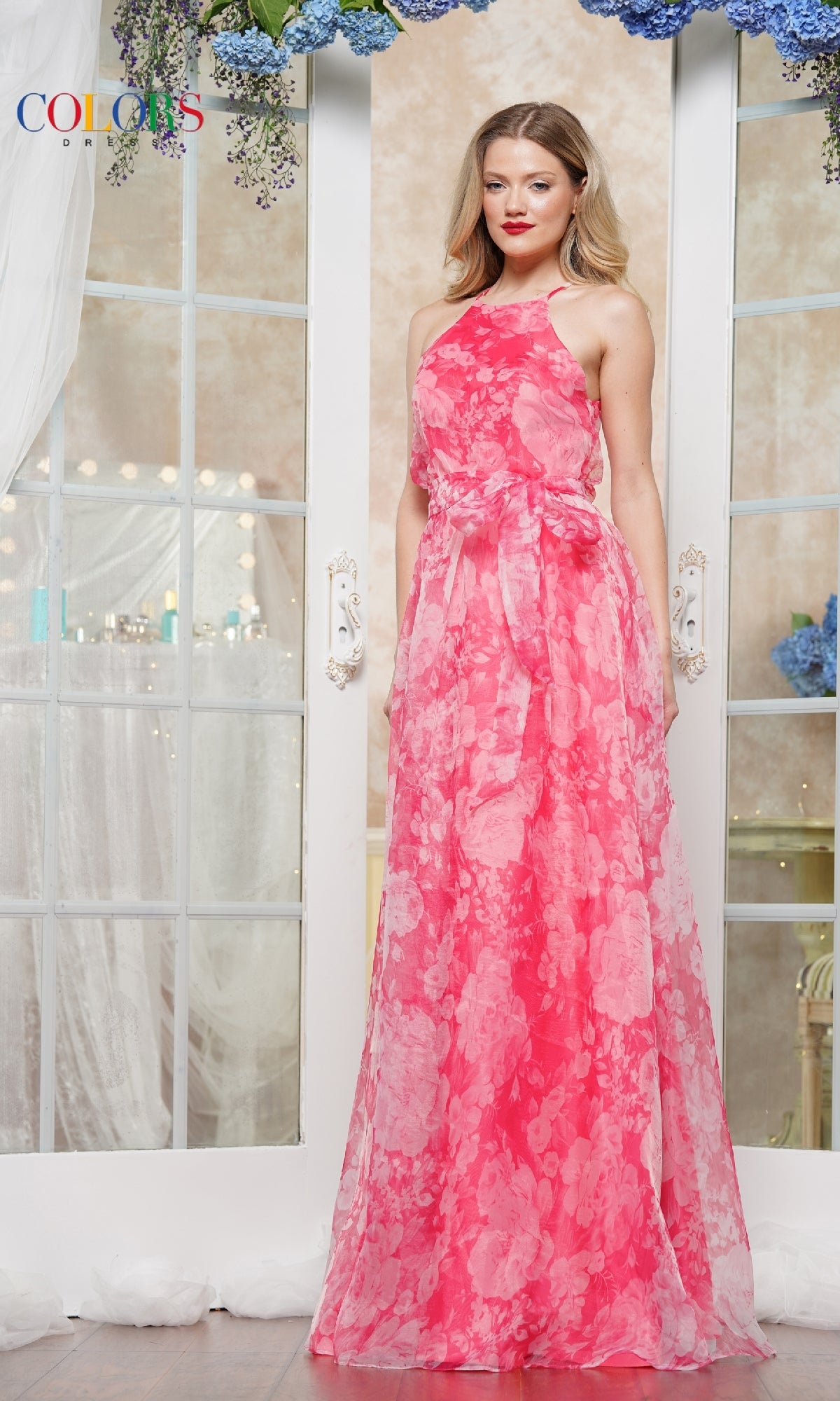 A woman stands indoors in the Colors Dress 3573, a long floral print prom gown. The sleeveless pink dress features a waist tie, with white doors and floral décor in the background.